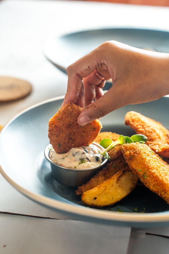 about-bg Hand dipping a crispy nugget into creamy sauce with potato wedges on a plate.