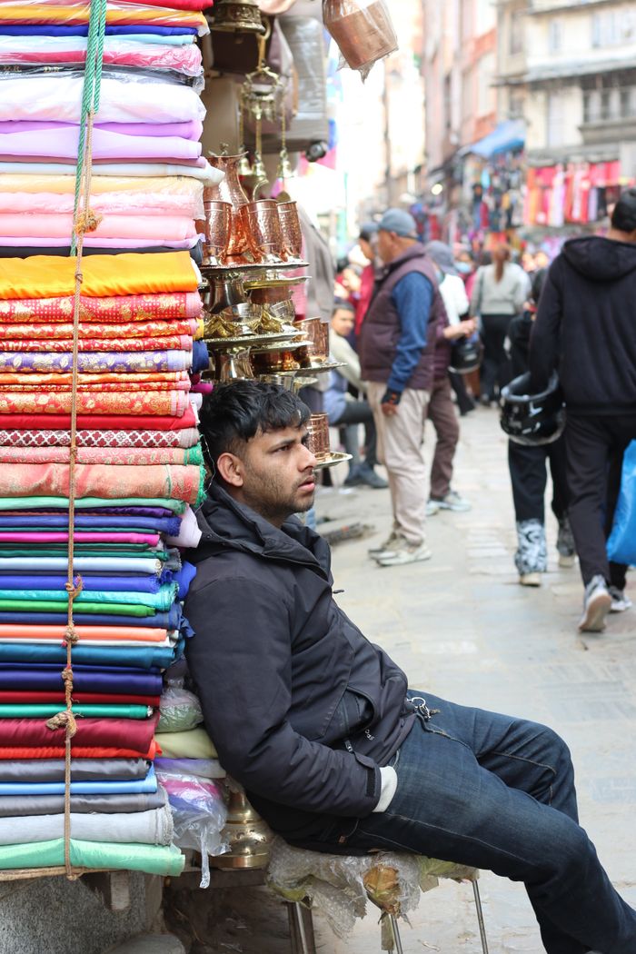 who-we-are A man sits beside vibrant fabrics and brass items on a Kathmandu street, capturing the bustling market life.