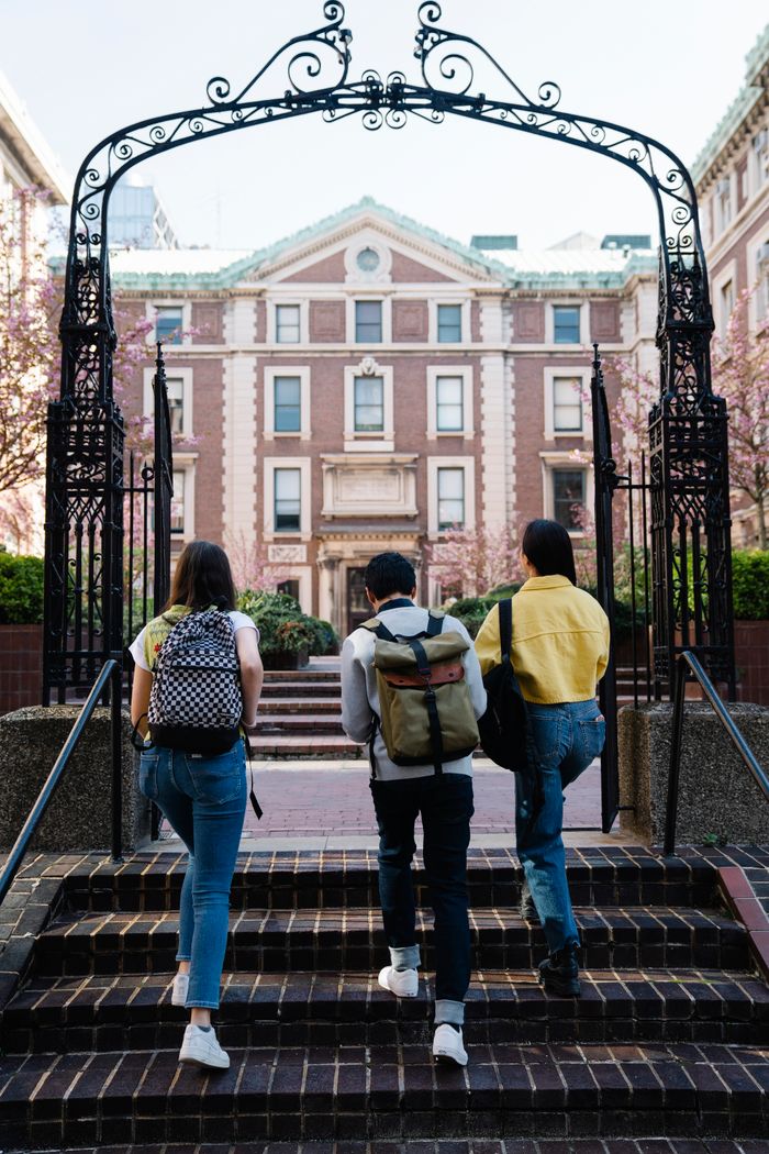 A group of students with backpacks walks towards a university building through an arch.